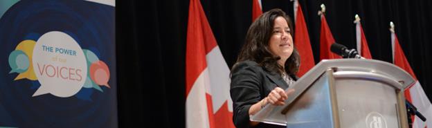 &Agrave; gauche, une banni&egrave;re avec plusieures bulles de parole avec l'une  que ce figure au plein milieu. Dans celle-ci est &eacute;crit "The Power of our  Voices". &Agrave; droite, la Ministre Jody Wilson-Raybould qui parle derri&egrave;re un  podium.